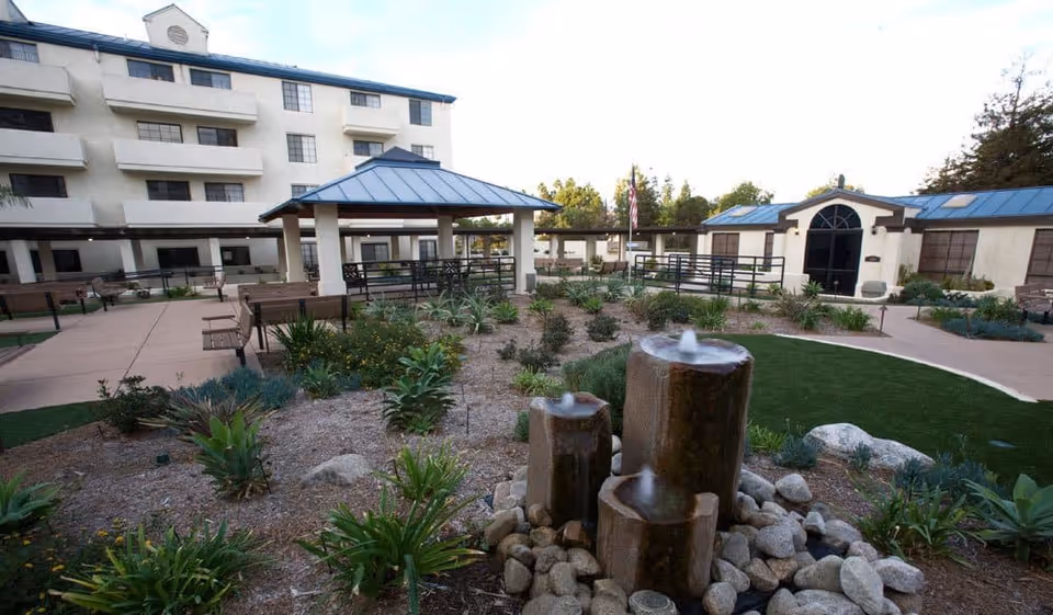 Outdoor courtyard area of a senior living facility with a three-tier stone water fountain surrounded by rocks and plants. There are paved walkways, benches, a covered seating area with a blue roof, and a multi-story building in the background. An American flag is visible near the center.