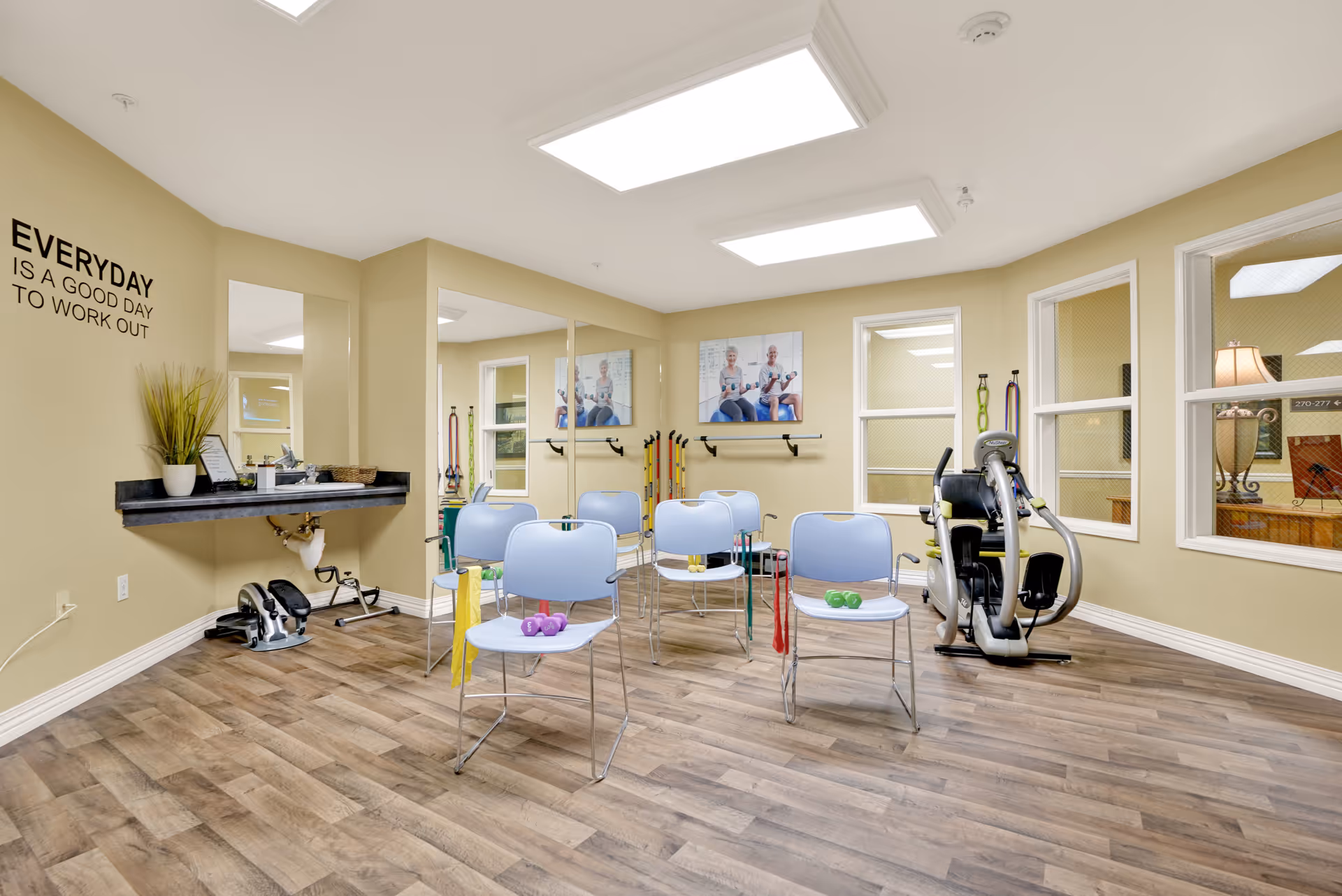 A bright exercise room with light wood flooring and beige walls. The room contains several light blue chairs arranged in rows, each with small exercise weights and resistance bands placed on or around them. There is a stationary exercise bike on the right side near three windows. A large mirror covers part of one wall, reflecting the room. On the left wall, there is a black floating shelf with a potted plant and some papers, and above it, a motivational quote reads 'EVERYDAY IS A GOOD DAY TO WORK OUT'. Two framed pictures of elderly people exercising hang on the back wall.