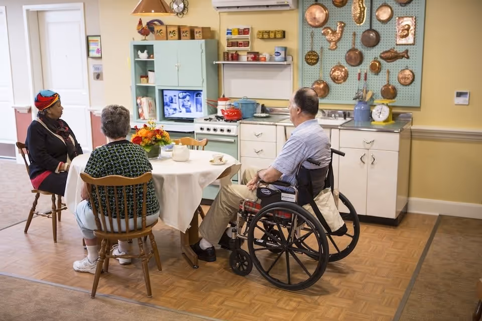 Three elderly individuals sitting around a round table with a white tablecloth in a cozy kitchen setting. One person is in a wheelchair, and the other two are seated on wooden chairs. The kitchen has vintage-style cabinets, a stove, and copper pots hanging on the wall. A small TV is on a shelf in the background.