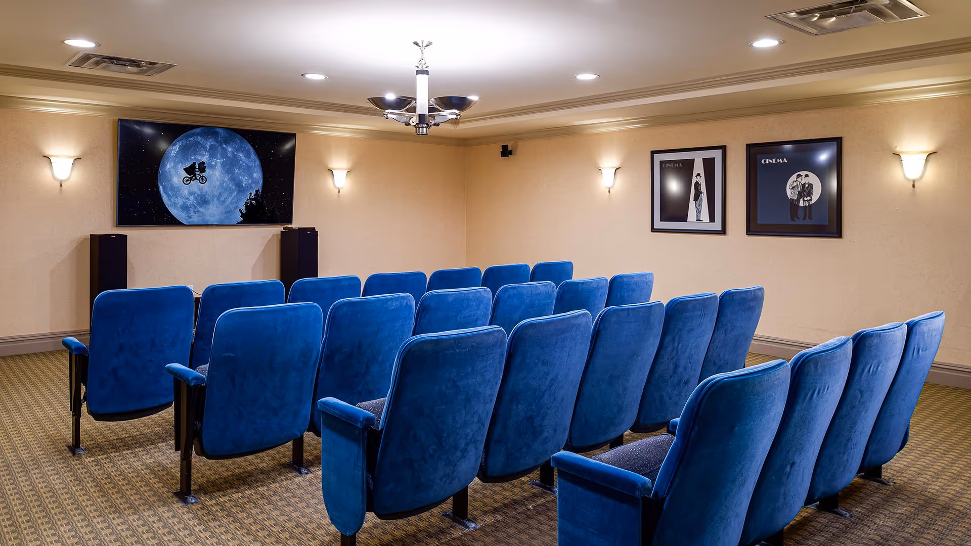 A small movie theater room with three rows of blue cushioned seats facing a large screen displaying an iconic scene of a bicycle flying in front of a full moon. The room has beige walls with wall sconces and two framed cinema-themed posters on the right wall.