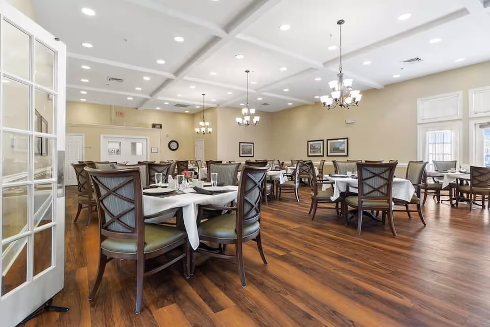 Bright, spacious dining room with multiple set tables and wooden chairs on hardwood floors under chandeliers.