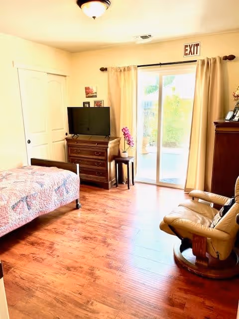 A bright senior living bedroom with wooden flooring, a bed with a patterned quilt, a wooden dresser with a flat-screen TV on top, a small side table with a purple orchid, a comfortable beige recliner chair, and sliding glass doors with beige curtains leading outside. An exit sign is visible above the sliding doors.