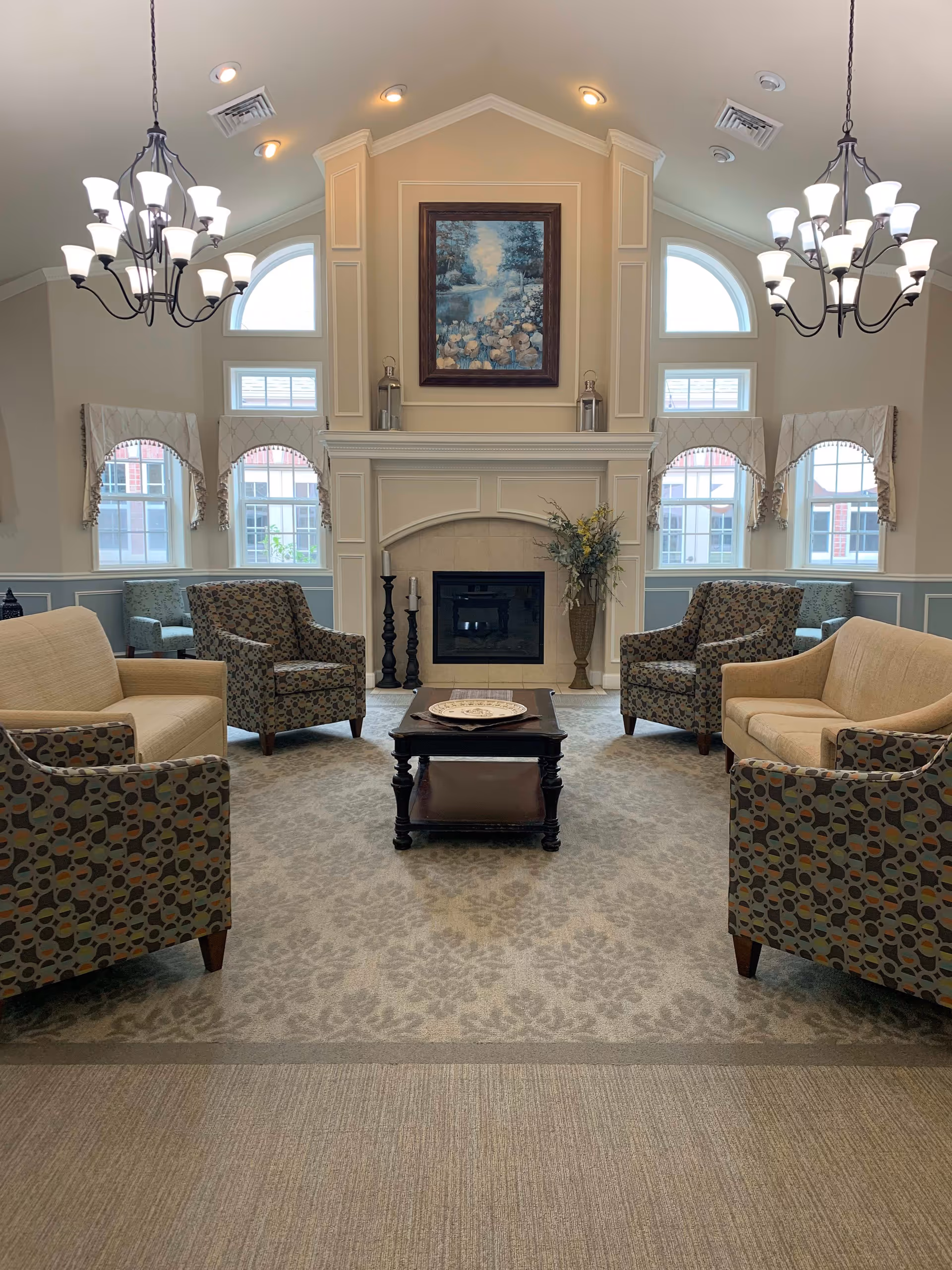 A cozy living room area in an assisted living facility featuring a fireplace with a framed painting above it, two chandeliers hanging from a vaulted ceiling, multiple windows with decorative valances, two beige sofas, four patterned armchairs, a dark wooden coffee table with a decorative plate, and a vase with flowers beside the fireplace.
