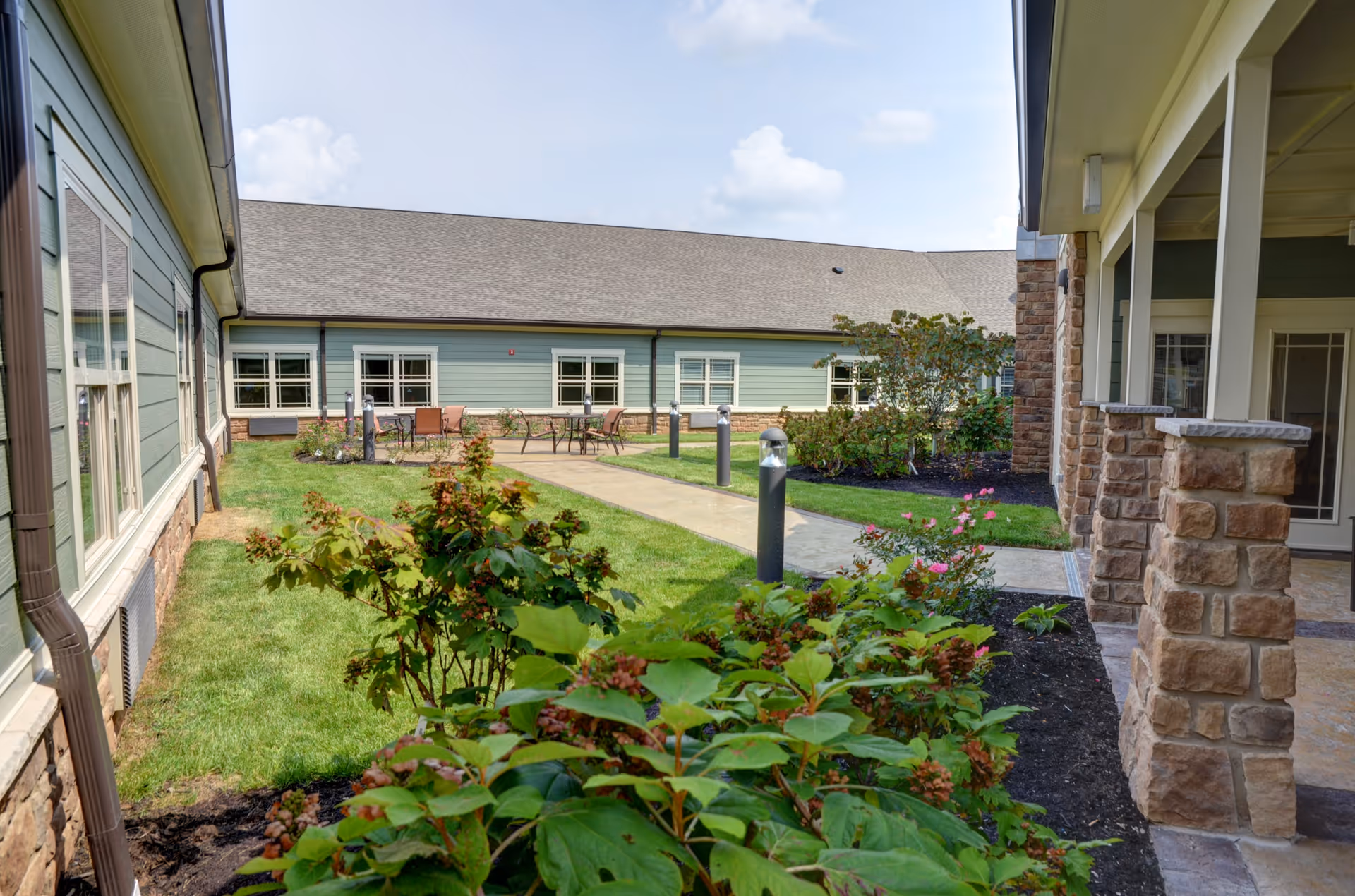 Outdoor courtyard area of a senior living facility with green grass, shrubs, and a paved walkway leading to a seating area with tables and chairs. The building has light green siding with stone accents and multiple windows under a clear sky.
