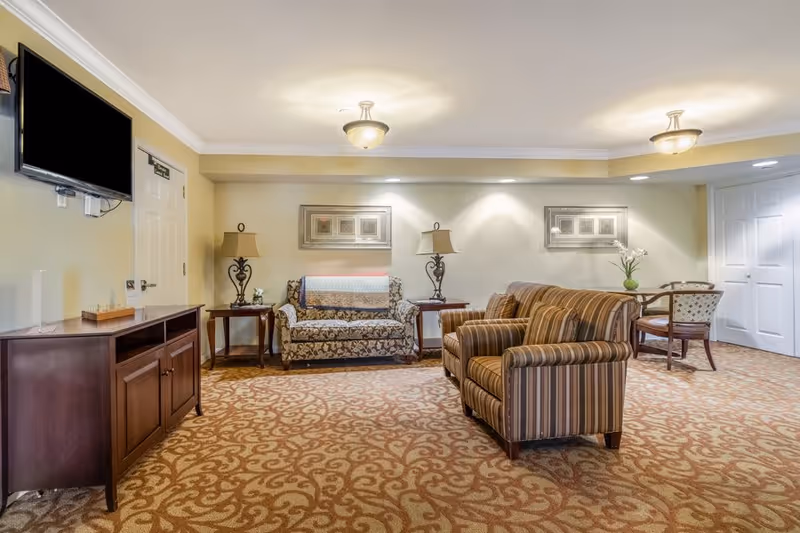 A cozy communal living room with patterned carpet, striped armchairs, a floral loveseat, side tables and lamps, wall art, a TV, and a small dining table.
