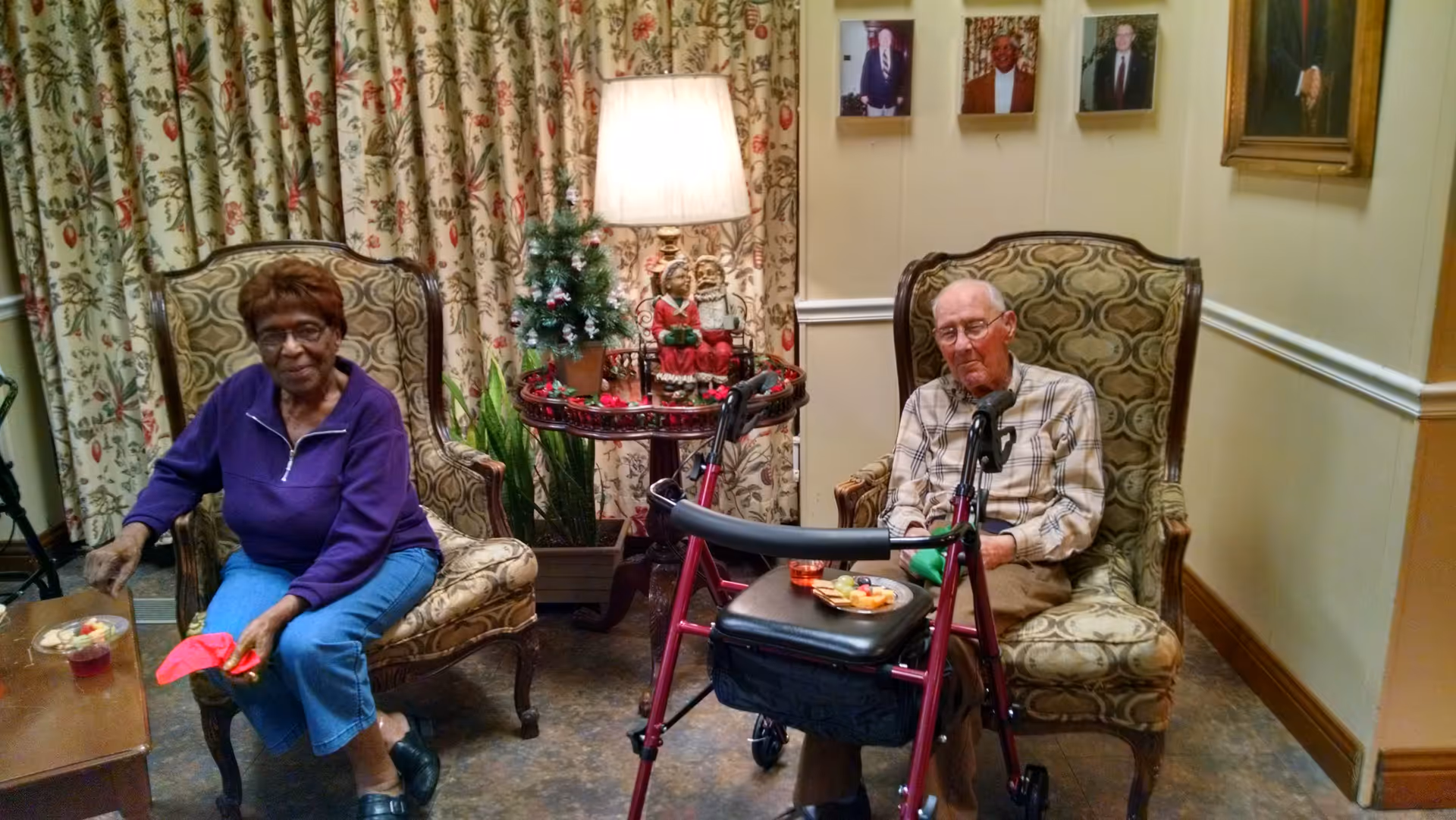 Two elderly individuals sitting in patterned armchairs in a cozy room with floral curtains and framed photos on the wall. A small table between them holds a lamp, a small decorated Christmas tree, and holiday figurines. A red walker is positioned in front of the man on the right, who is wearing a plaid shirt and glasses. The woman on the left is wearing a purple top and blue pants, holding a red napkin. Both have small plates of snacks and drinks nearby.