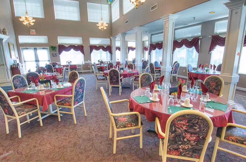 A spacious dining room with multiple round and square tables covered with red tablecloths. Each table is set with green placemats, glassware, napkins folded upright, and small flower arrangements. The room features large windows with maroon valances, high ceilings with chandeliers, and carpeted floors. White columns separate sections of the room.