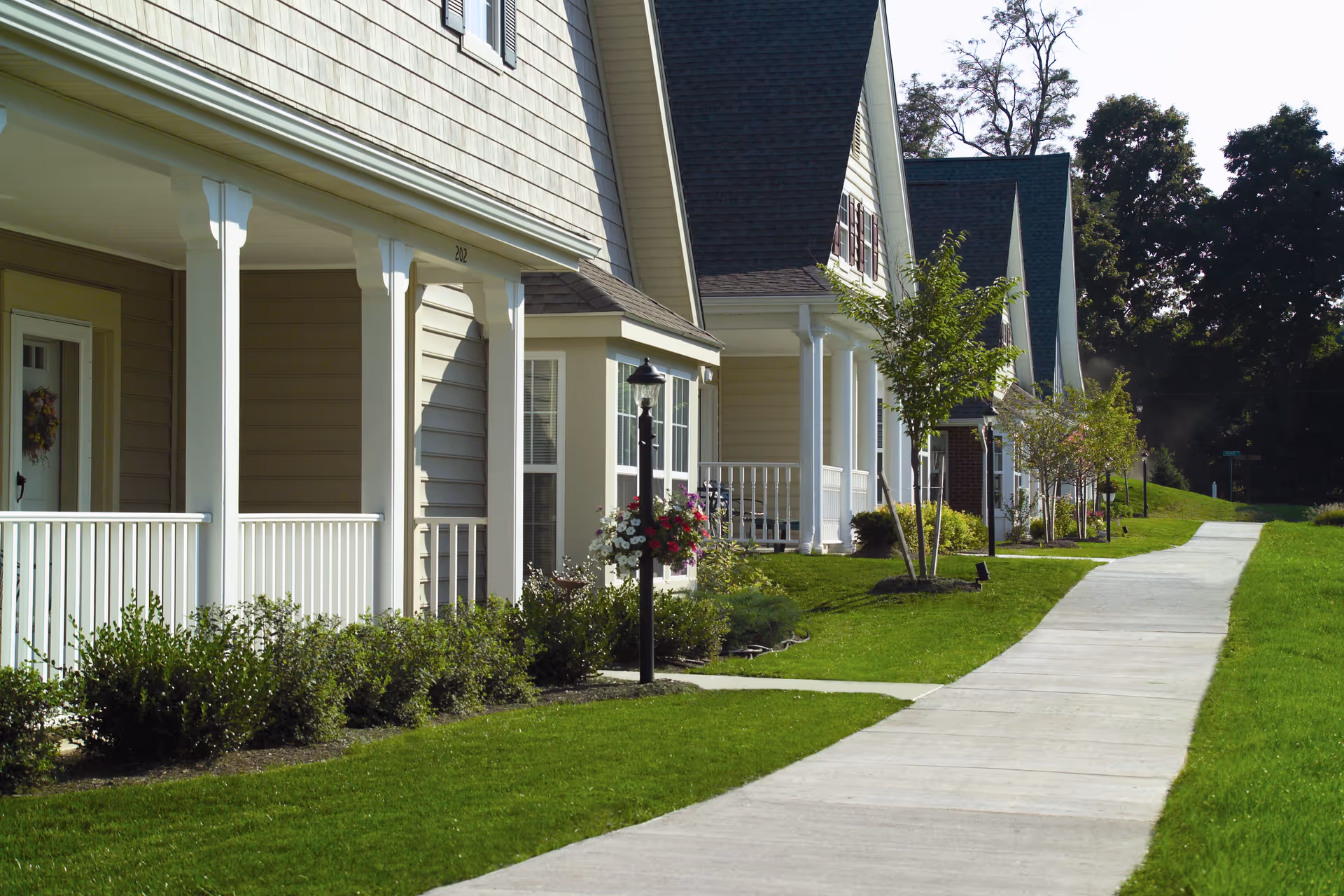 A row of beige residential buildings with white trim and porches, lined with small bushes and trees along a concrete sidewalk on a sunny day.