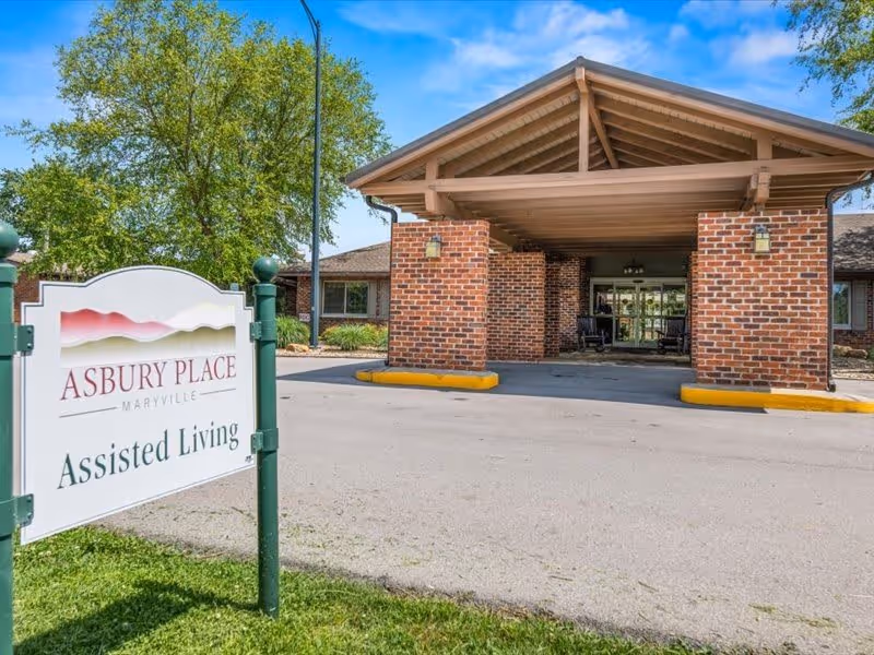 Entrance of Asbury Place Maryville assisted living facility showing a covered drop-off area with brick pillars and a sign in the foreground on a grassy patch that reads 'Asbury Place Maryville Assisted Living'.