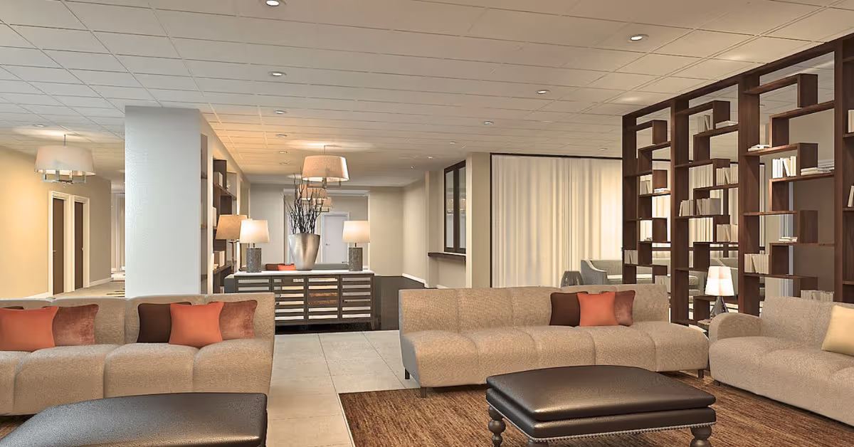 A spacious and well-lit living room area in a senior living facility featuring beige sofas with orange and brown cushions, a dark brown ottoman, a large wooden bookshelf filled with books, and a console table with lamps and decorative items. The room has a tiled floor, recessed ceiling lights, and a neutral color palette.