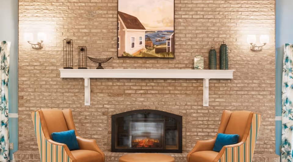 A cozy living room area with a brick fireplace in the center. Above the fireplace is a white mantel decorated with vases and metal sculptures, and a painting of a coastal house scene hangs on the brick wall. Two striped armchairs with blue cushions face each other with a round ottoman between them. Wall-mounted light fixtures are on either side of the fireplace, and floral curtains frame the sides of the image.