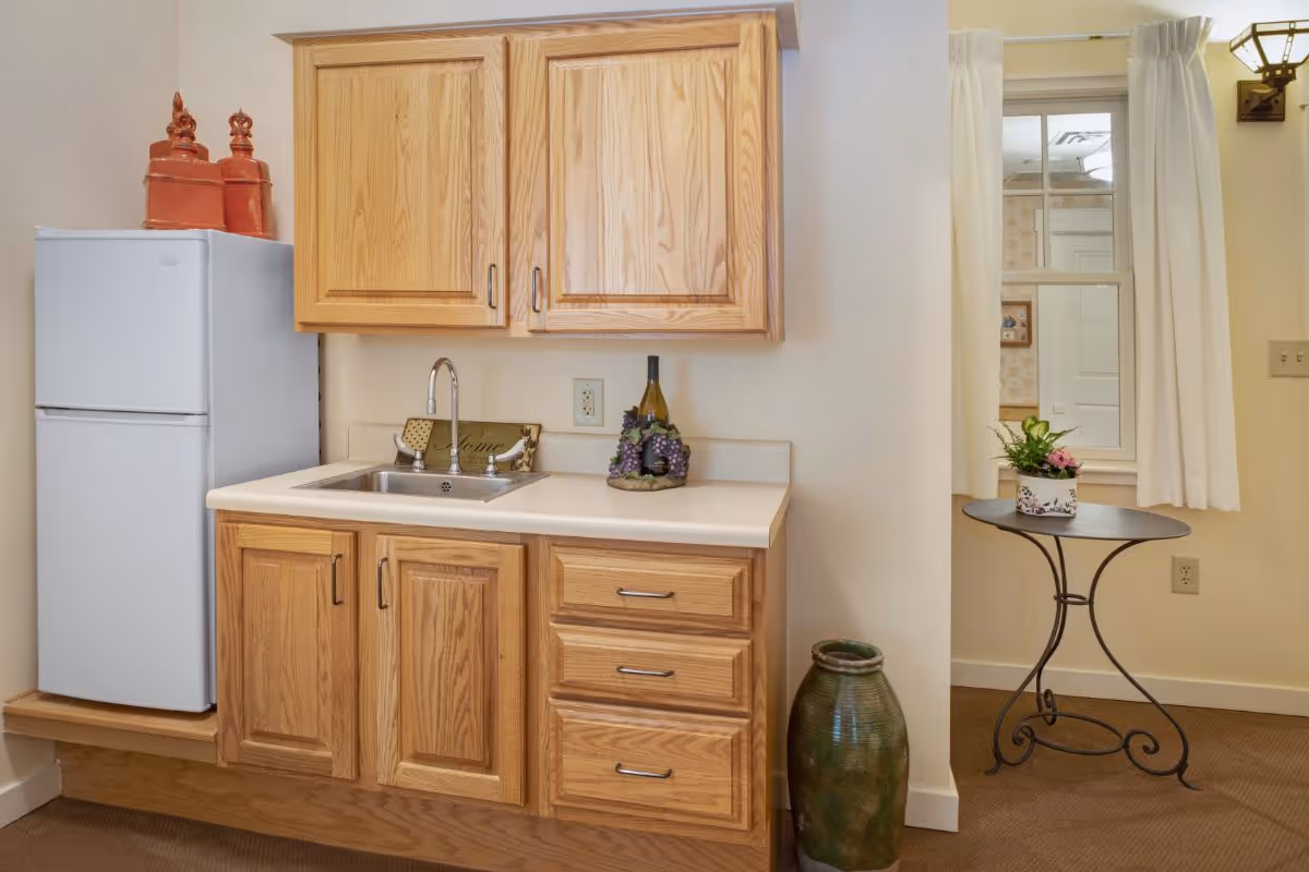 A small kitchen area with wooden cabinets, a white refrigerator, a sink, and a countertop decorated with a wine bottle and grape ornament. To the right, there is a small round table with a potted plant in front of a window with white curtains.