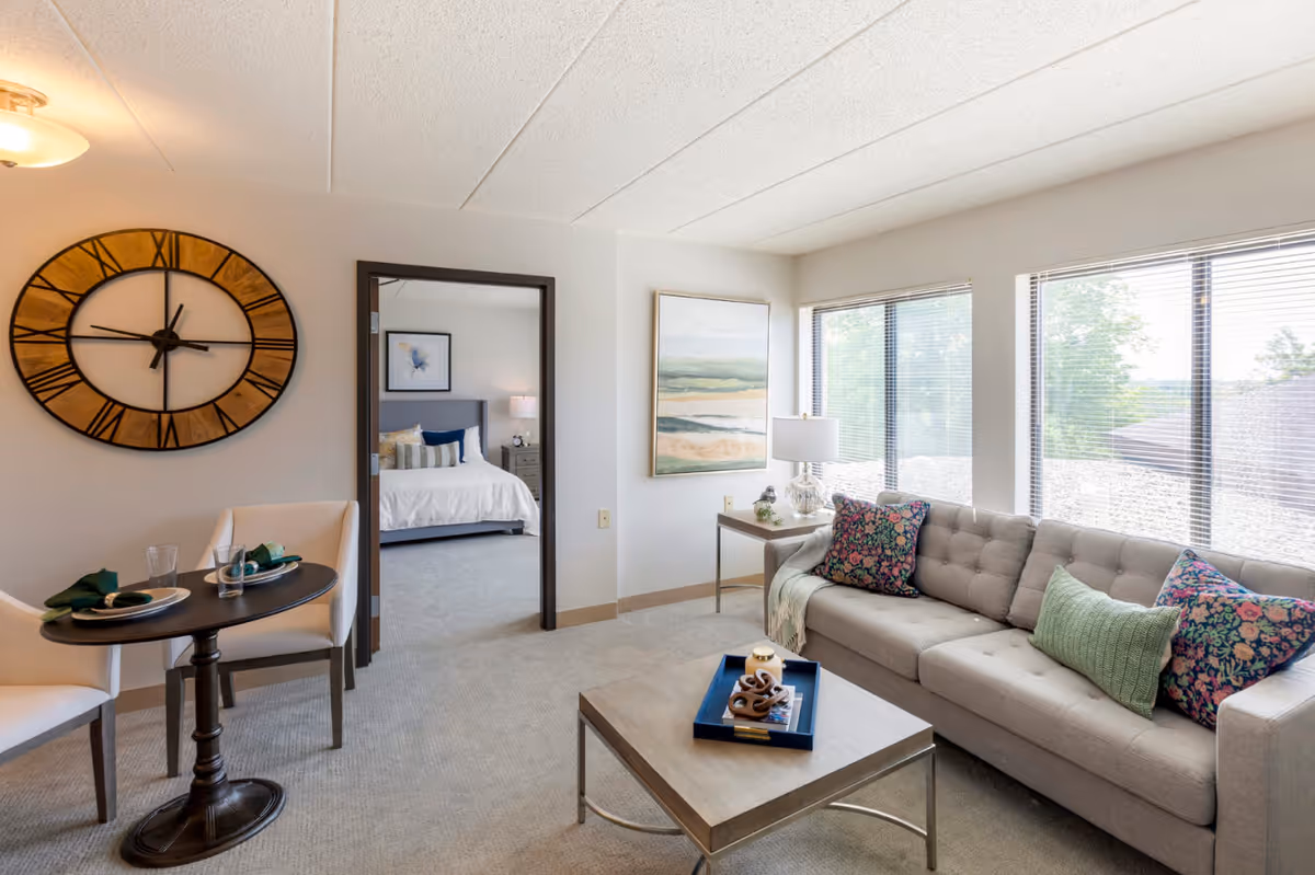Bright furnished living room with a sofa, coffee table, small dining table and a doorway leading to a bedroom.