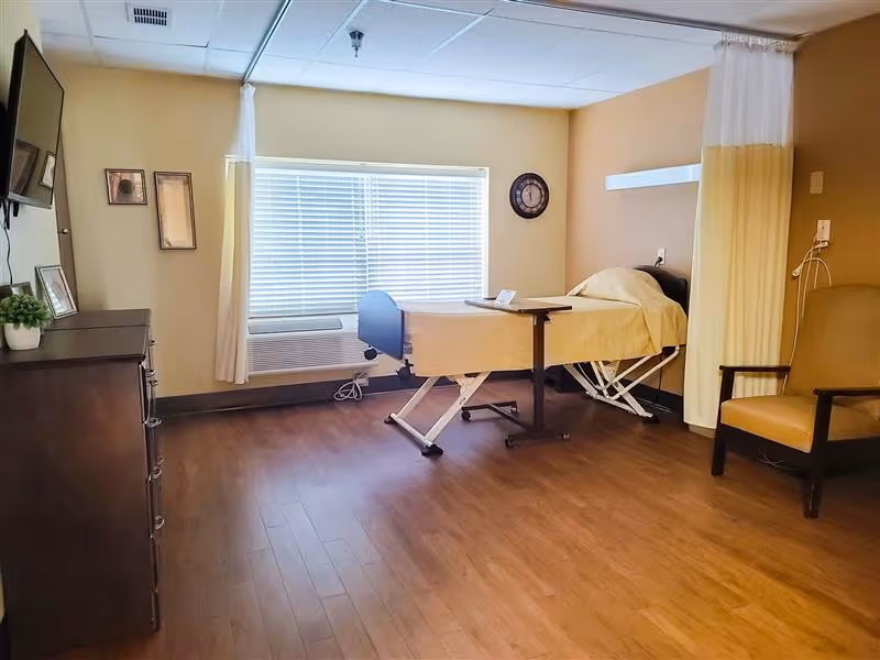 A nursing facility room with a hospital bed covered in a beige blanket, a wooden dresser with a small plant and picture frames, a wall-mounted TV, a large window with blinds, a clock on the wall, a beige armchair, and a privacy curtain partially drawn.