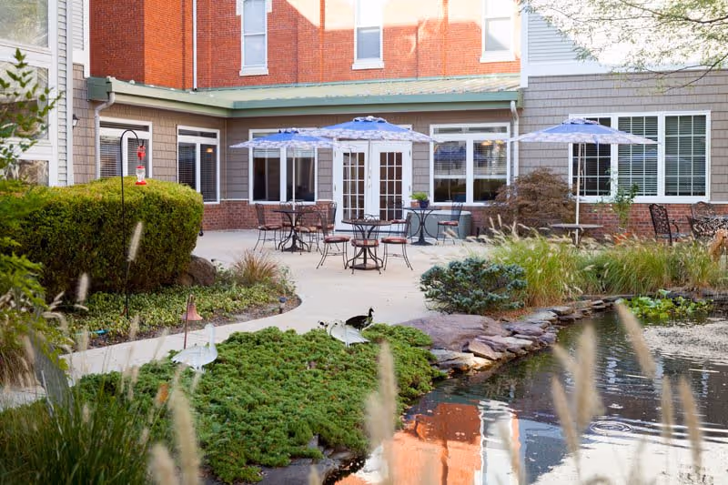 Outdoor patio area at Woodbury Mews with several tables and chairs under blue umbrellas, surrounded by greenery and a small pond with rocks in the foreground. The building exterior features brick and siding with multiple windows and French doors leading to the patio.