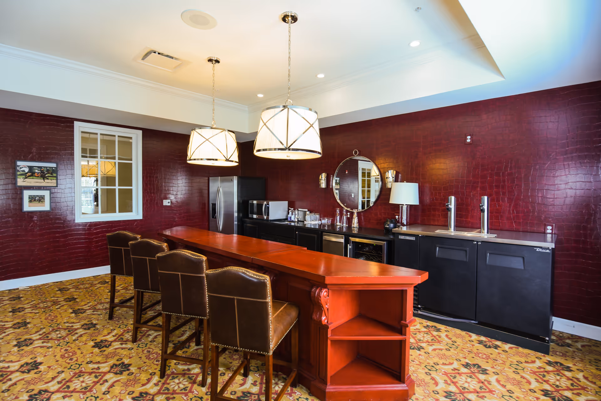 Interior view of a room with a long wooden counter and four brown leather chairs. The back wall is dark red with a textured pattern, featuring a round mirror, a lamp, and two beverage taps on a black cabinet. Two pendant lights hang from the ceiling, and the floor is covered with a patterned carpet.