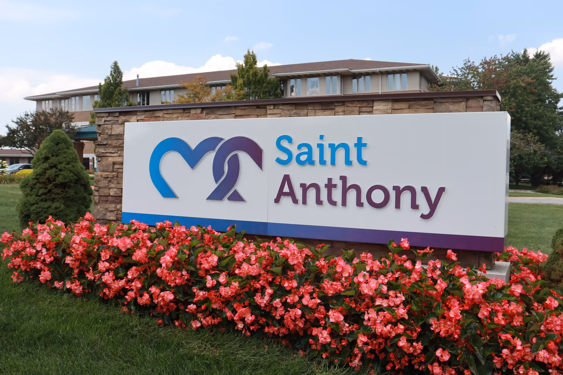 Outdoor view of a sign for Saint Anthony facility with a stone wall behind it, surrounded by green grass and vibrant pink flowers, with a multi-story building and trees in the background under a partly cloudy sky.