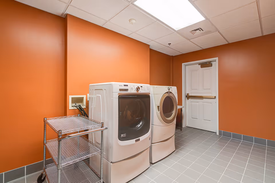 Laundry room with two front-loading machines, a metal shelving cart, orange walls and tiled floor.