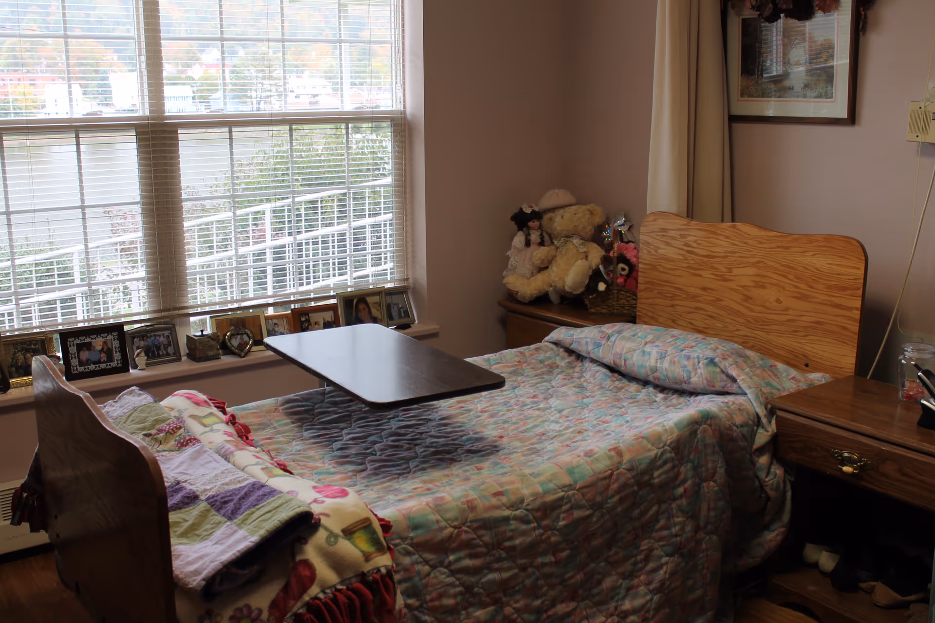 A cozy bedroom in a healthcare facility featuring a single wooden bed with a colorful quilt and a small overbed table. The room has a large window with blinds, a windowsill lined with framed photographs, a nightstand with stuffed animals, and a wooden desk with various items on it.