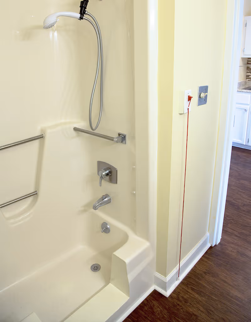 A beige bathtub and shower combination with a handheld showerhead, two horizontal grab bars, and a built-in seat. There is a red emergency pull cord on the wall next to the tub, and a view into an adjacent room with wood flooring and white cabinetry.