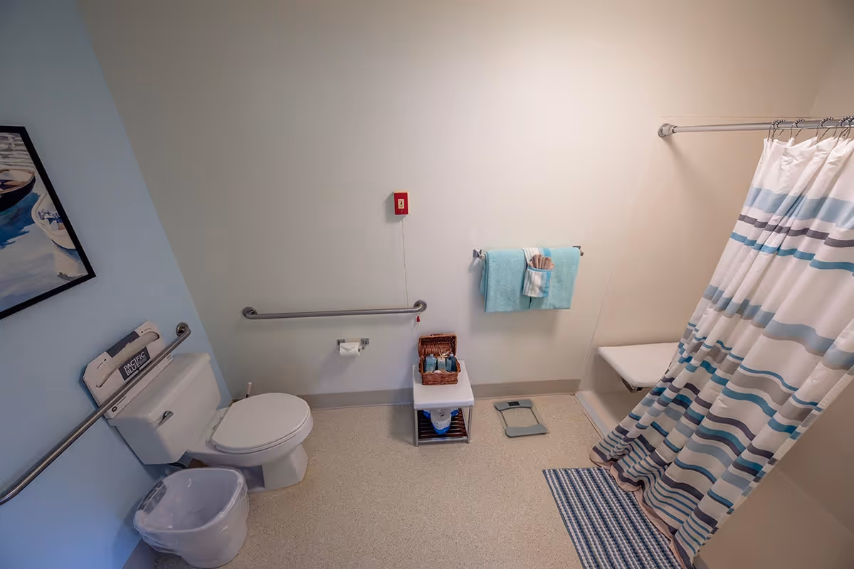 A clean and accessible bathroom with a toilet, grab bars on the walls, a small white table holding toiletries, a towel rack with folded blue towels, a striped shower curtain, a shower bench, a floor scale, and a blue and white striped bath mat. There is also a trash can next to the toilet and a framed painting on the light blue wall.