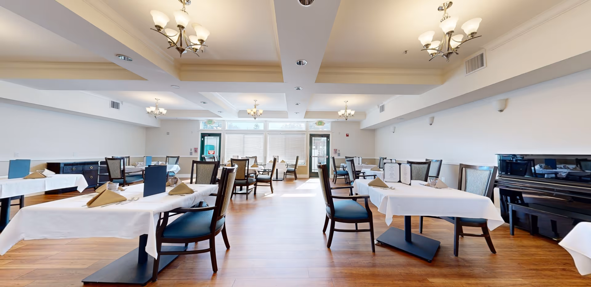 A spacious dining room with multiple tables covered in white tablecloths, each set with folded napkins and menus. The room features wooden flooring, several chandeliers hanging from a coffered ceiling, and large windows letting in natural light. There is a black piano against the right wall.