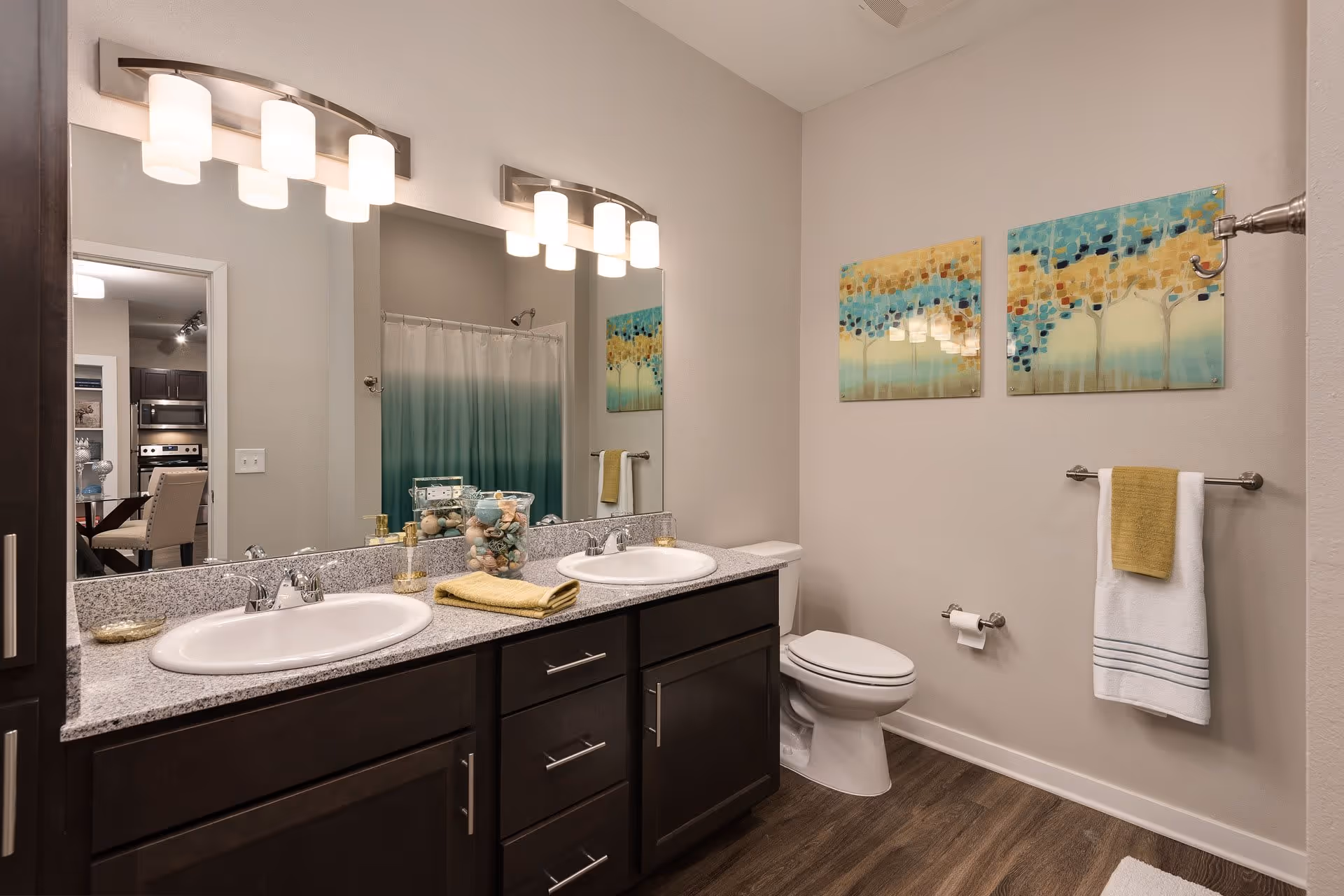 Modern bathroom with double sinks set in a granite countertop, dark wood cabinets, a toilet, and a shower with a blue-green ombre curtain. Two colorful abstract paintings hang on the wall above a towel rack with a white and yellow towel. The bathroom has wood flooring and bright overhead lighting.