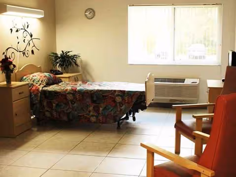 A senior facility bedroom with a bed, dressers, plants, and a large window.