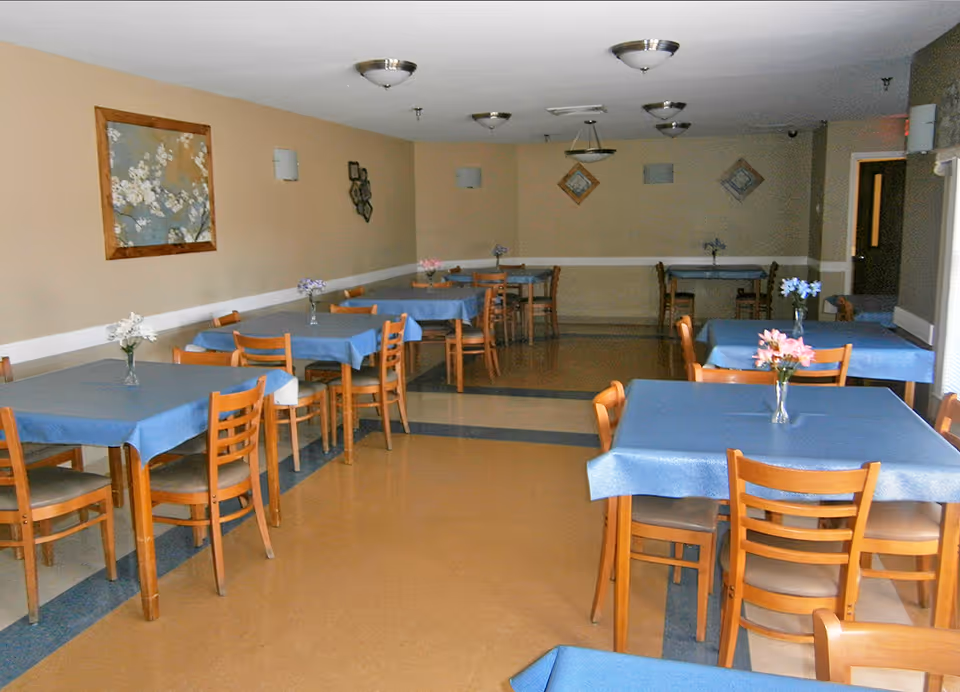 A dining room with multiple wooden tables covered with blue tablecloths, each table decorated with a small vase of flowers. The room has beige walls with framed artwork and several ceiling light fixtures. Wooden chairs are arranged around the tables, and there is a door visible at the far end of the room.