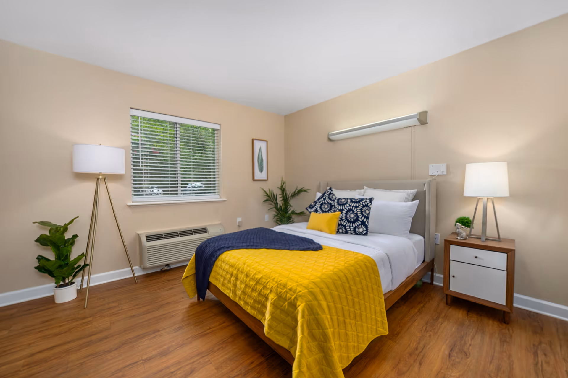 A neatly made bed with a yellow quilt and blue throw blanket in a senior living bedroom. The bed has multiple pillows including blue patterned and white ones. There is a wooden nightstand with a lamp and small plant on the right side of the bed. A tall floor lamp and a potted plant are on the left side near a window with white blinds. The room has beige walls and wood flooring.