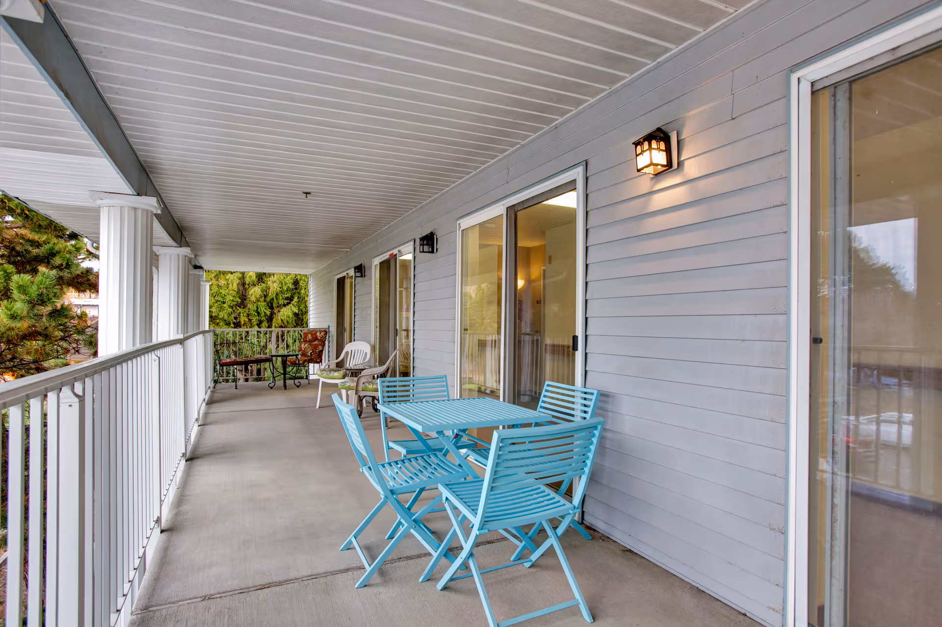 Covered outdoor balcony area with light blue metal table and four matching chairs, white railing, white columns, and additional seating with a small table in the background. The balcony is attached to a building with sliding glass doors and exterior wall lights.