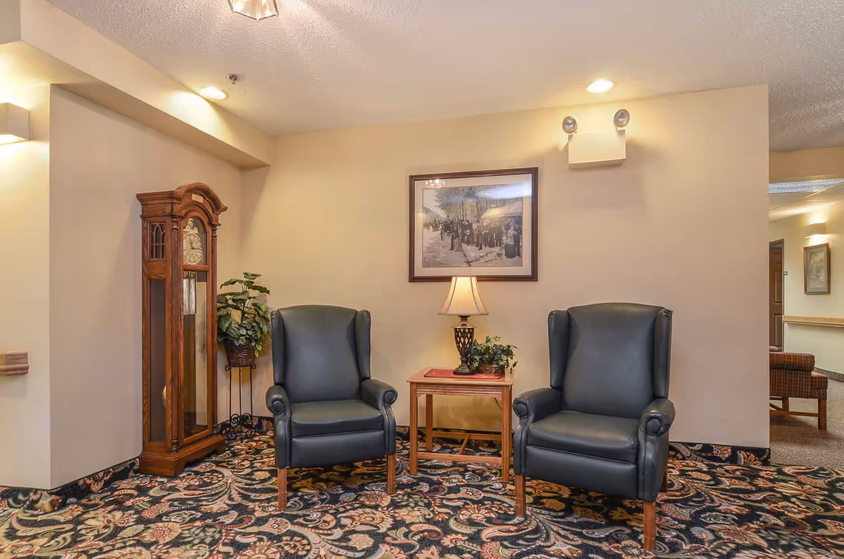 A cozy sitting area in a senior living facility with two dark green leather armchairs separated by a wooden side table holding a lamp and a small plant. Behind the chairs is a framed picture on a beige wall. To the left, there is a tall wooden grandfather clock and a potted plant on a stand. The floor is covered with a patterned carpet in dark colors.