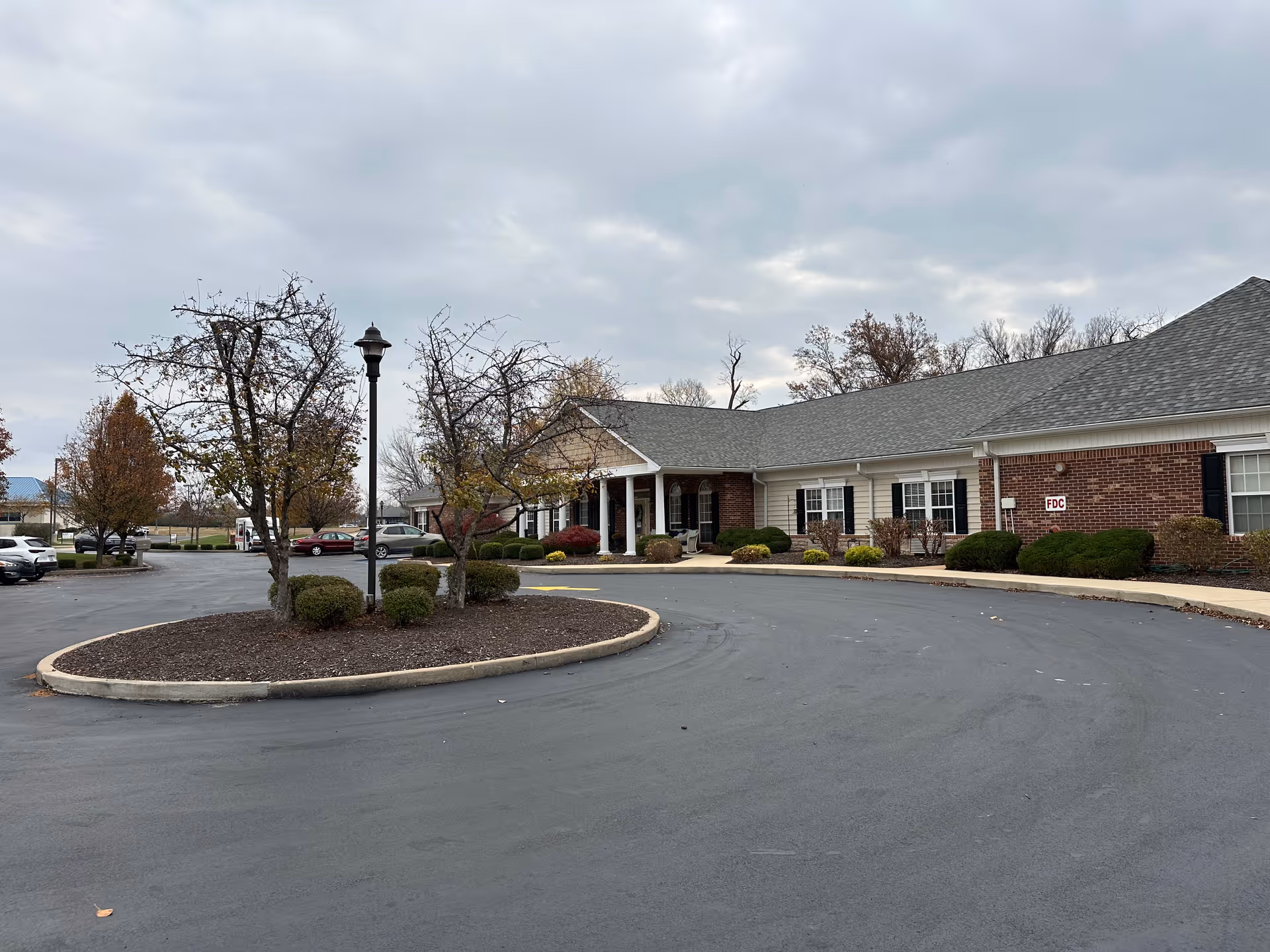 Exterior view of a single-story senior living facility building with a curved driveway and landscaped island with small trees and bushes. Several parked cars are visible in the background under a cloudy sky.
