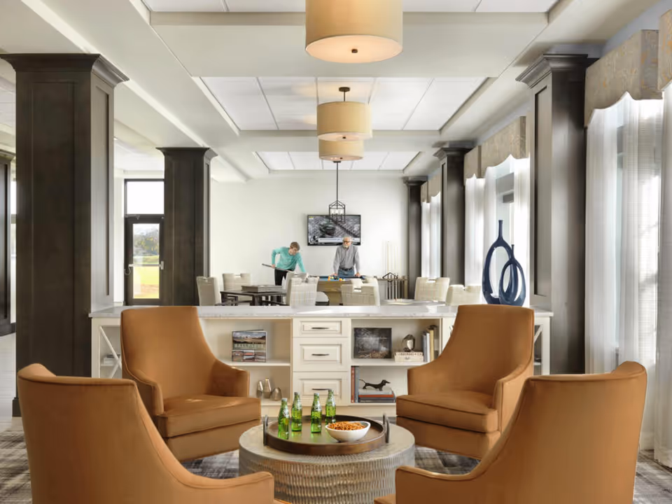 A bright and spacious common area in a senior living facility featuring four brown armchairs arranged around a round coffee table with green glass bottles and a bowl of snacks. Behind the seating area is a white counter with shelves holding books and decorative items. In the background, two people are playing a game at a table, with large windows and dark pillars lining the room. The ceiling has recessed lighting and hanging lamps.