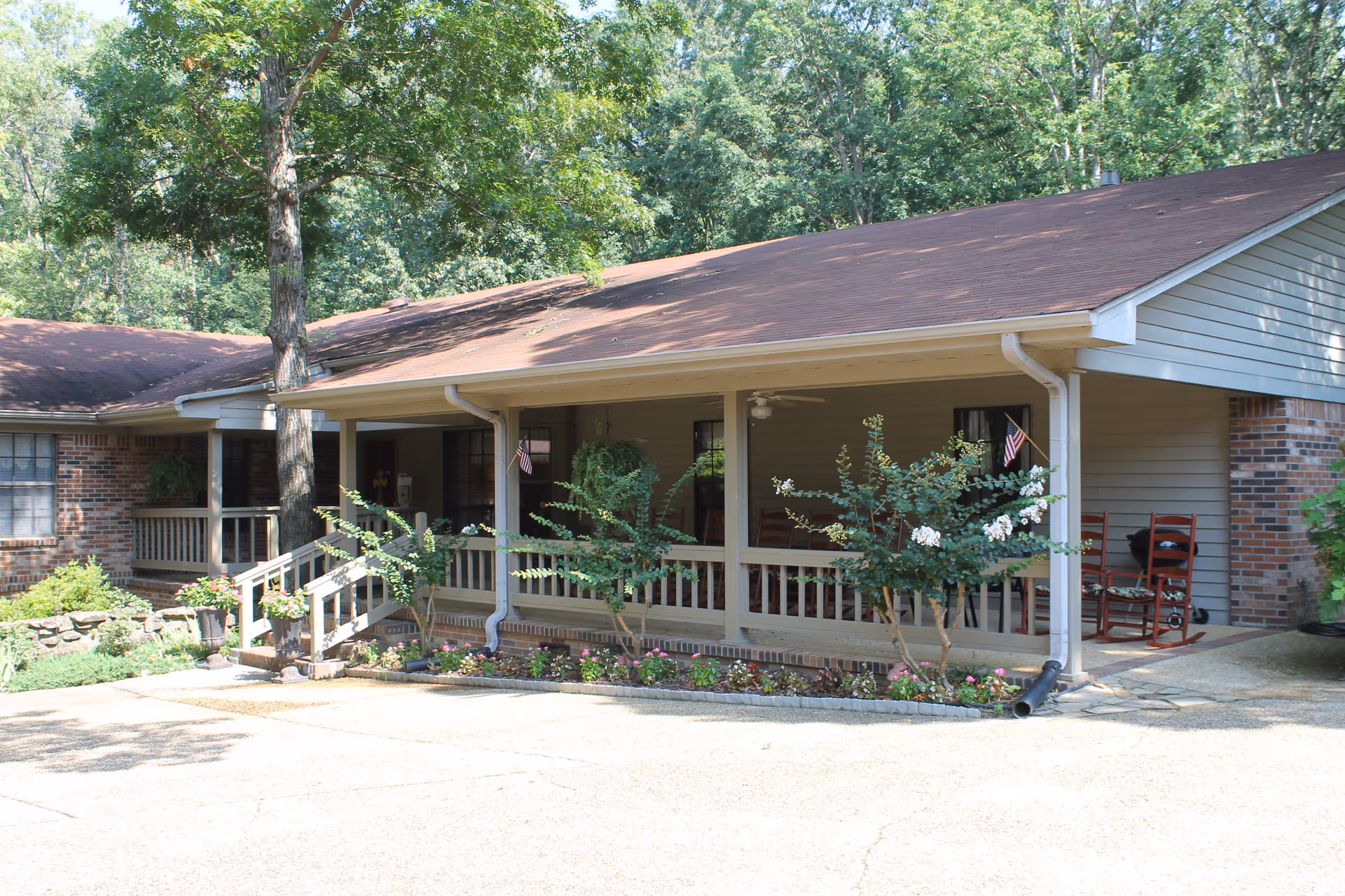 Exterior view of a single-story residential building with a covered porch. The porch has railings, rocking chairs, small American flags, and is surrounded by greenery and flower beds. The building has a brown roof and a mix of brick and siding walls, with trees in the background.