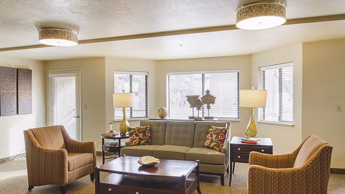 Bright furnished living room with a sofa, two patterned armchairs, a coffee table and side tables with lamps in front of windows.