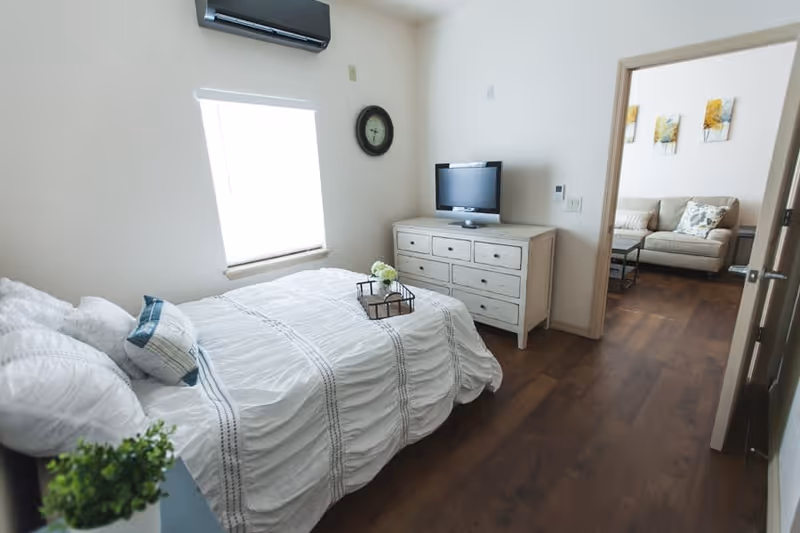 A bright bedroom with a neatly made bed covered in white bedding with decorative pillows. A small tray with flowers is placed on the bed. There is a white dresser with a flat-screen TV on top against the wall. A window with a blind and a wall clock are above the dresser. The room has wooden flooring and an air conditioning unit mounted above the window. Through an open door, a living room with a beige sofa and wall art is visible.