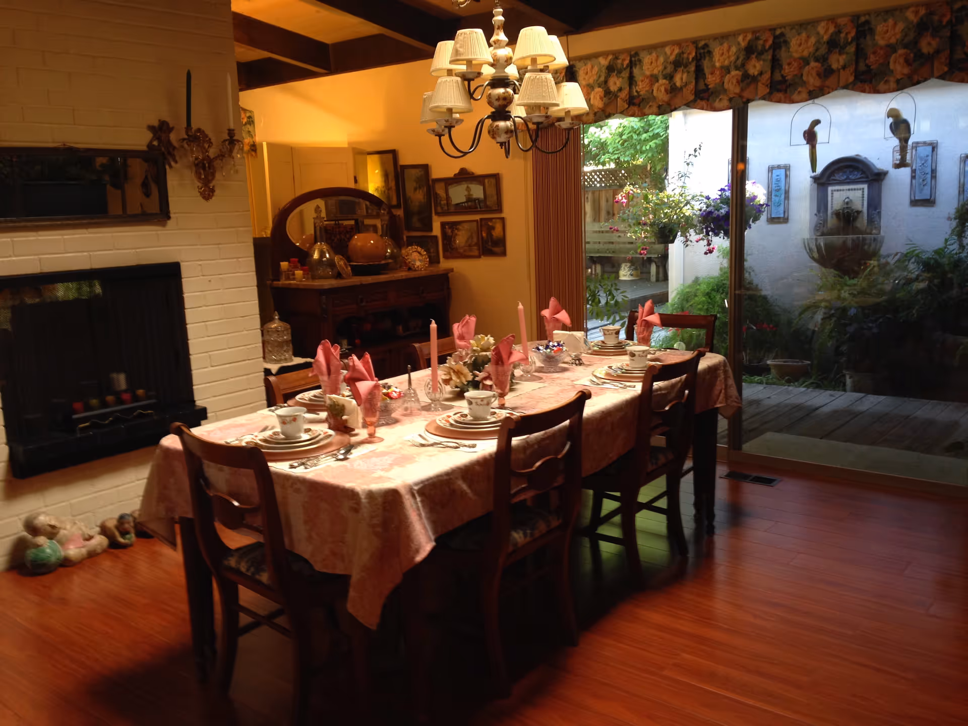 A warmly lit dining room with a wooden table set for six people, featuring pink napkins, candles, and fine china. The room has wooden floors, a white brick fireplace on the left, and a large sliding glass door on the right that opens to a garden patio with plants and a wall fountain.
