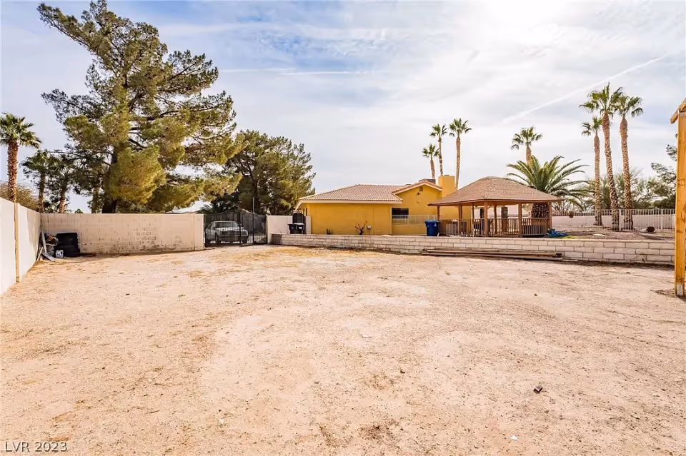 A large, open dirt yard area enclosed by a low concrete block wall with a yellow building and a wooden gazebo structure in the background. Tall palm trees and other trees surround the yard under a partly cloudy sky.