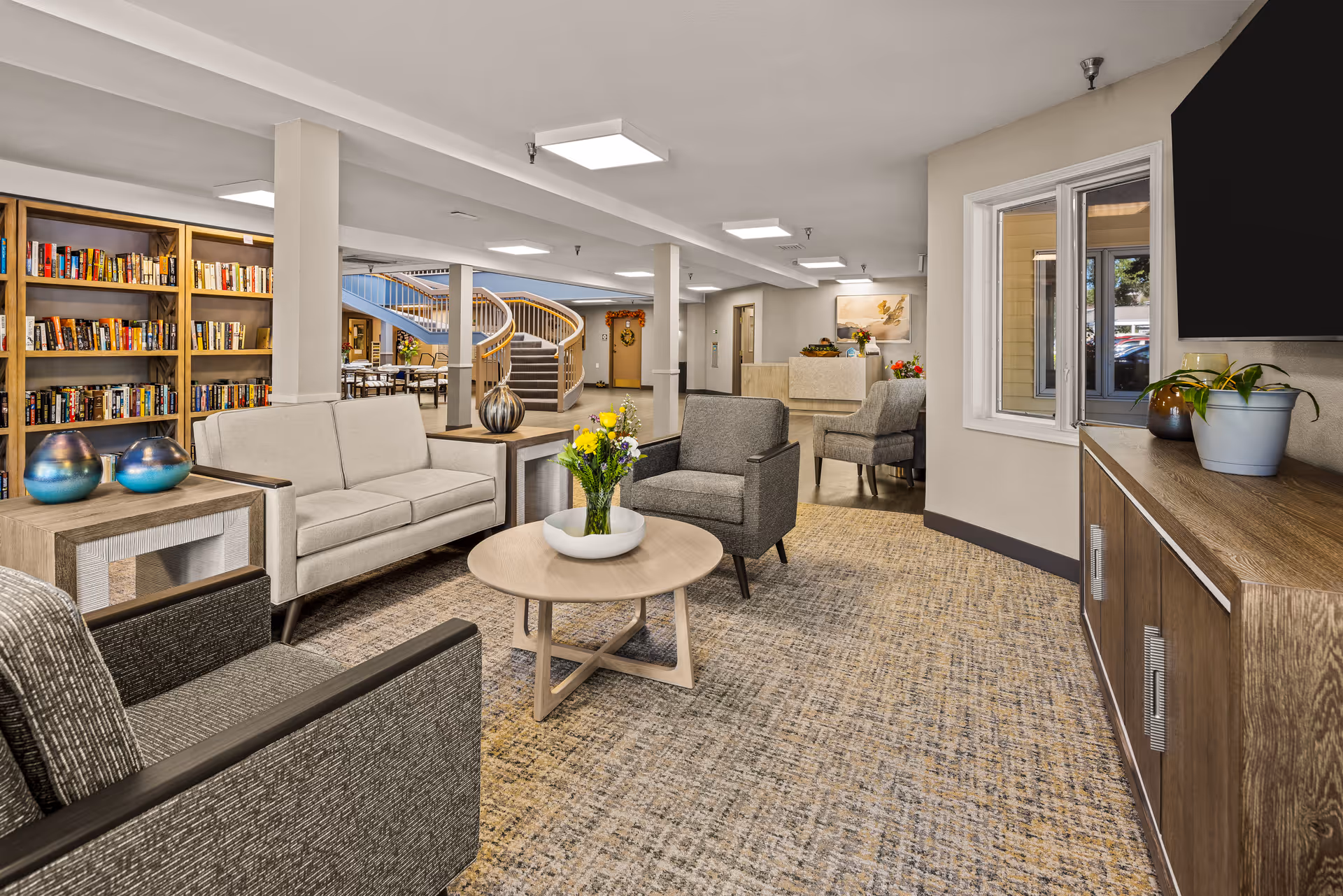 A spacious senior living community lounge area featuring comfortable seating with a beige sofa, two gray armchairs, and a round wooden coffee table with a flower arrangement. In the background, there is a bookshelf filled with books, a curved staircase, and a reception desk. The room has neutral-colored walls, carpeted floors, and modern ceiling lights.