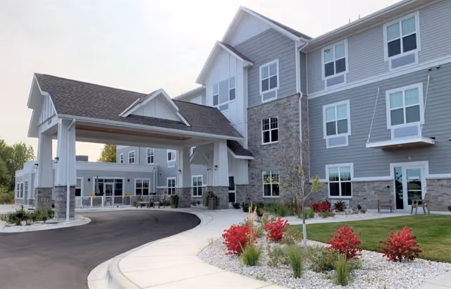 Exterior view of a modern senior living facility building with gray siding and stone accents. The building has multiple windows and a covered entrance driveway. There are landscaped areas with red shrubs and green grass along a curved sidewalk leading to the entrance.
