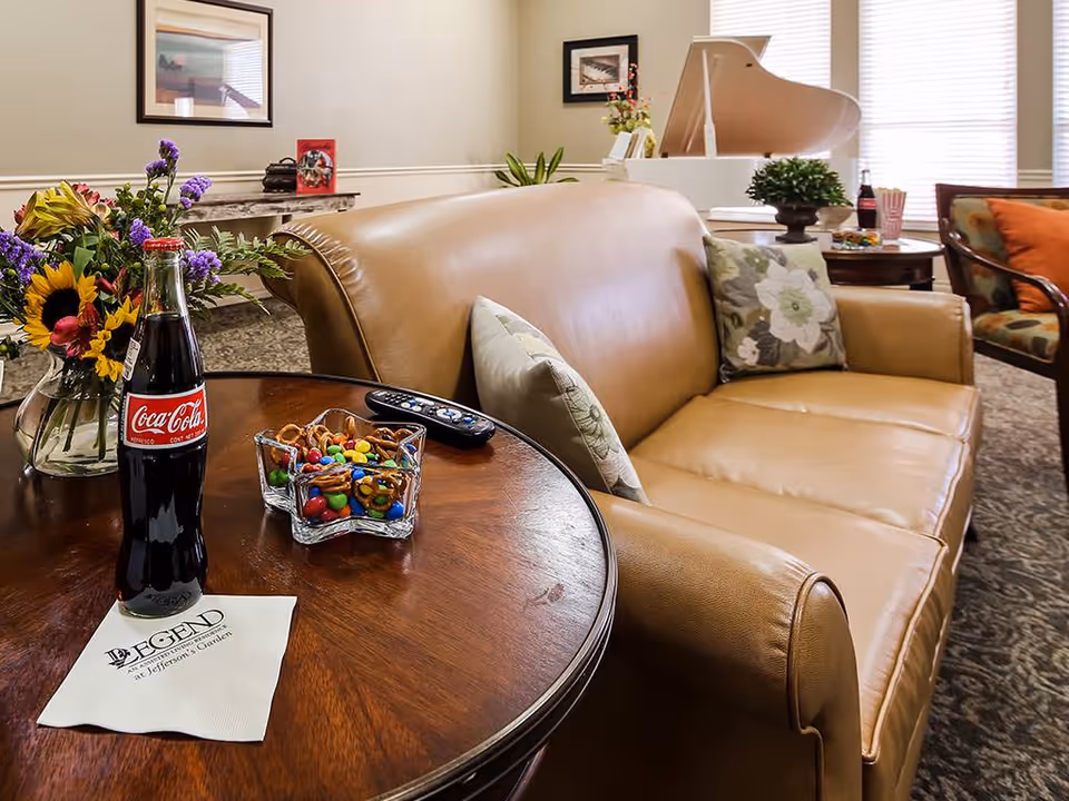 A cozy living room area with a tan leather sofa adorned with floral pillows, a round wooden table holding a glass bottle of Coca-Cola, a bowl of pretzels and colorful candies, a TV remote, and a vase of fresh flowers. In the background, there is a white grand piano near a window with blinds, a patterned armchair with an orange pillow, and framed artwork on the walls.