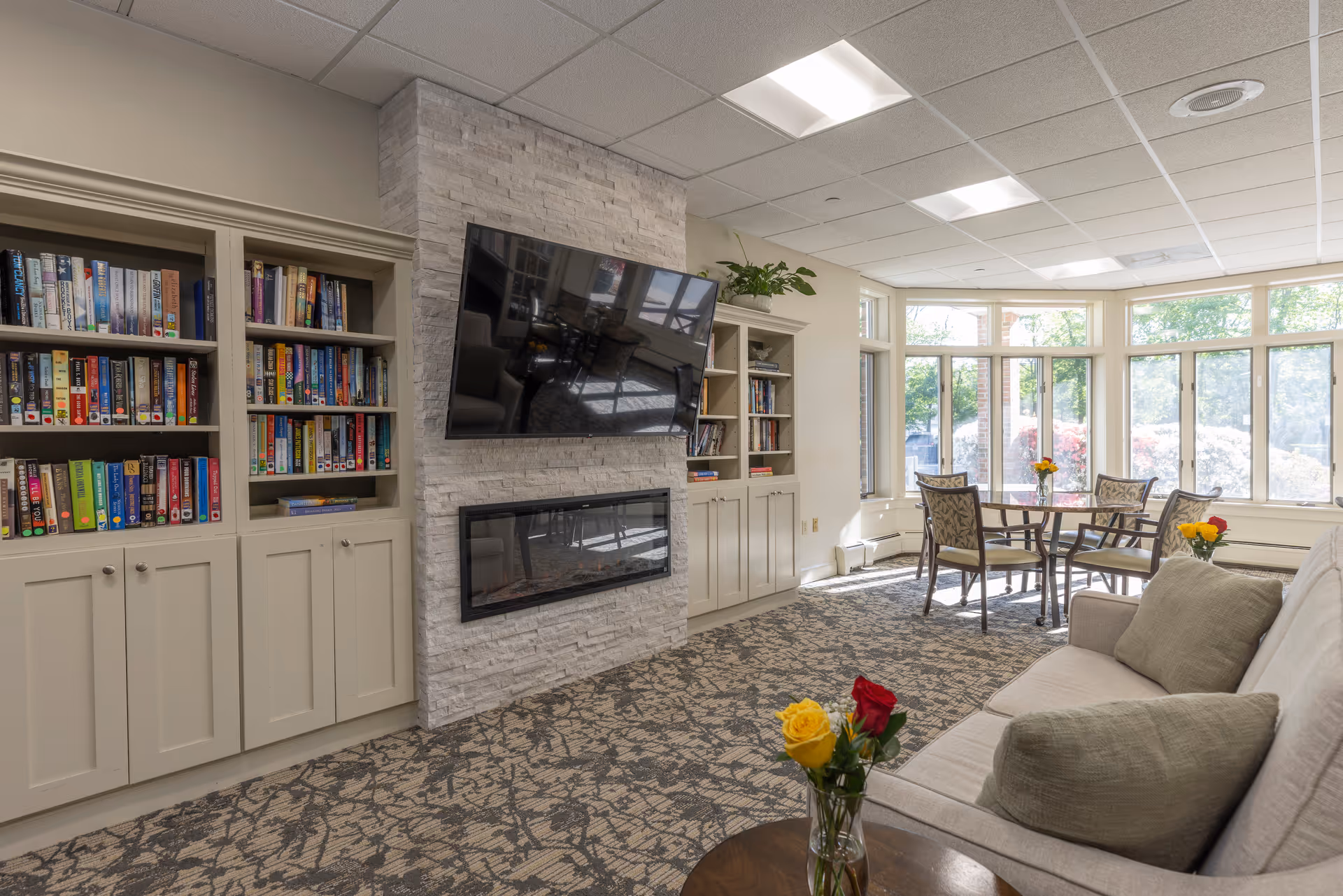 A bright and cozy living room area with a beige sofa adorned with cushions, a round wooden table with a vase of yellow and red roses, a wall-mounted flat-screen TV above a modern fireplace set in a stone accent wall, built-in bookshelves filled with books, and a dining table with four chairs near large windows letting in natural light.