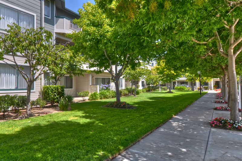 A well-maintained outdoor garden area at a senior living facility with green grass, trees providing shade, flower beds along a concrete walkway, and the exterior of the building with windows and balconies visible.