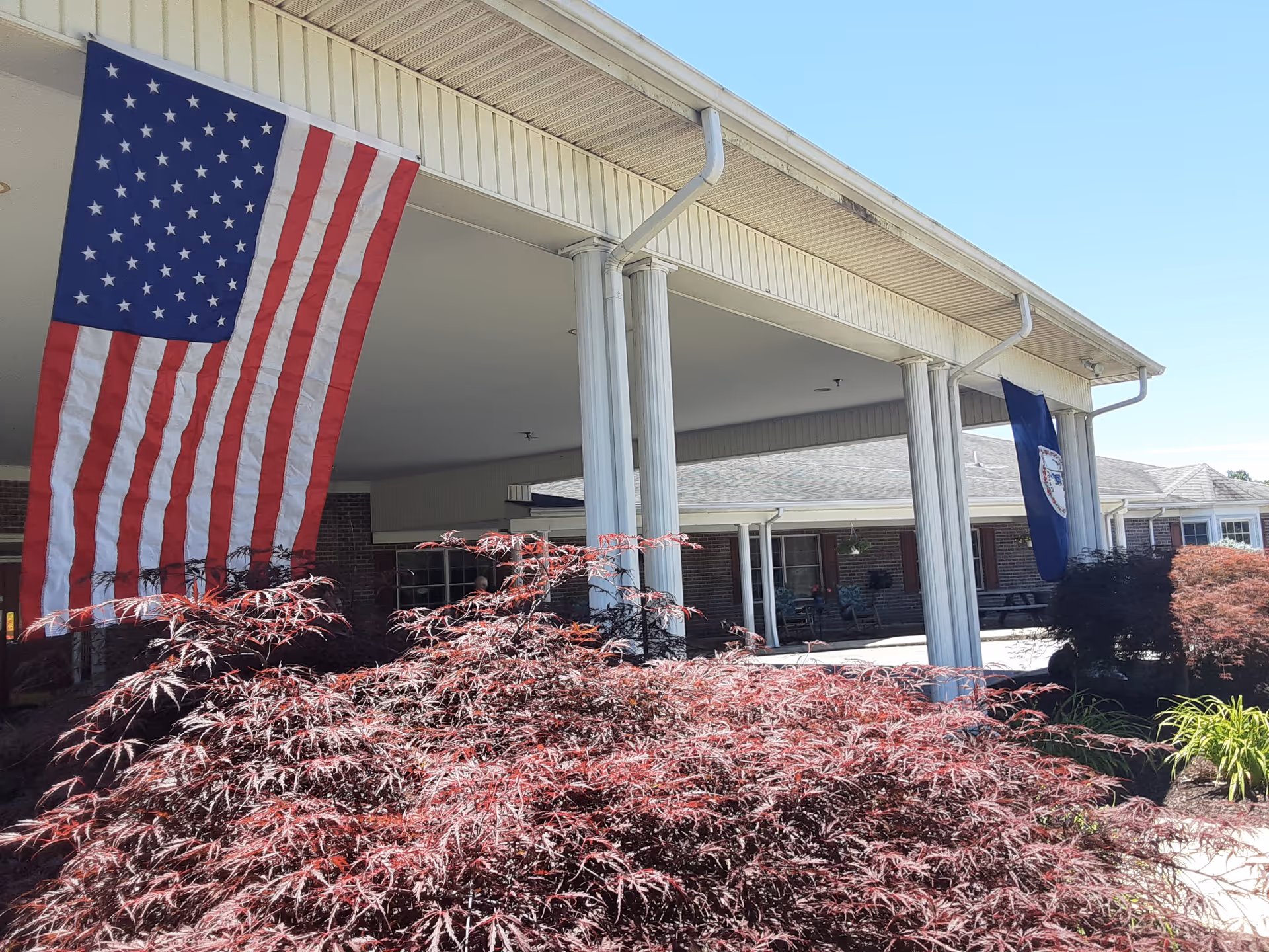 Exterior view of Birch Gardens Assisted Living showing a covered entrance with white columns, an American flag and another flag hanging from the roof, and red foliage in the foreground under a clear blue sky.