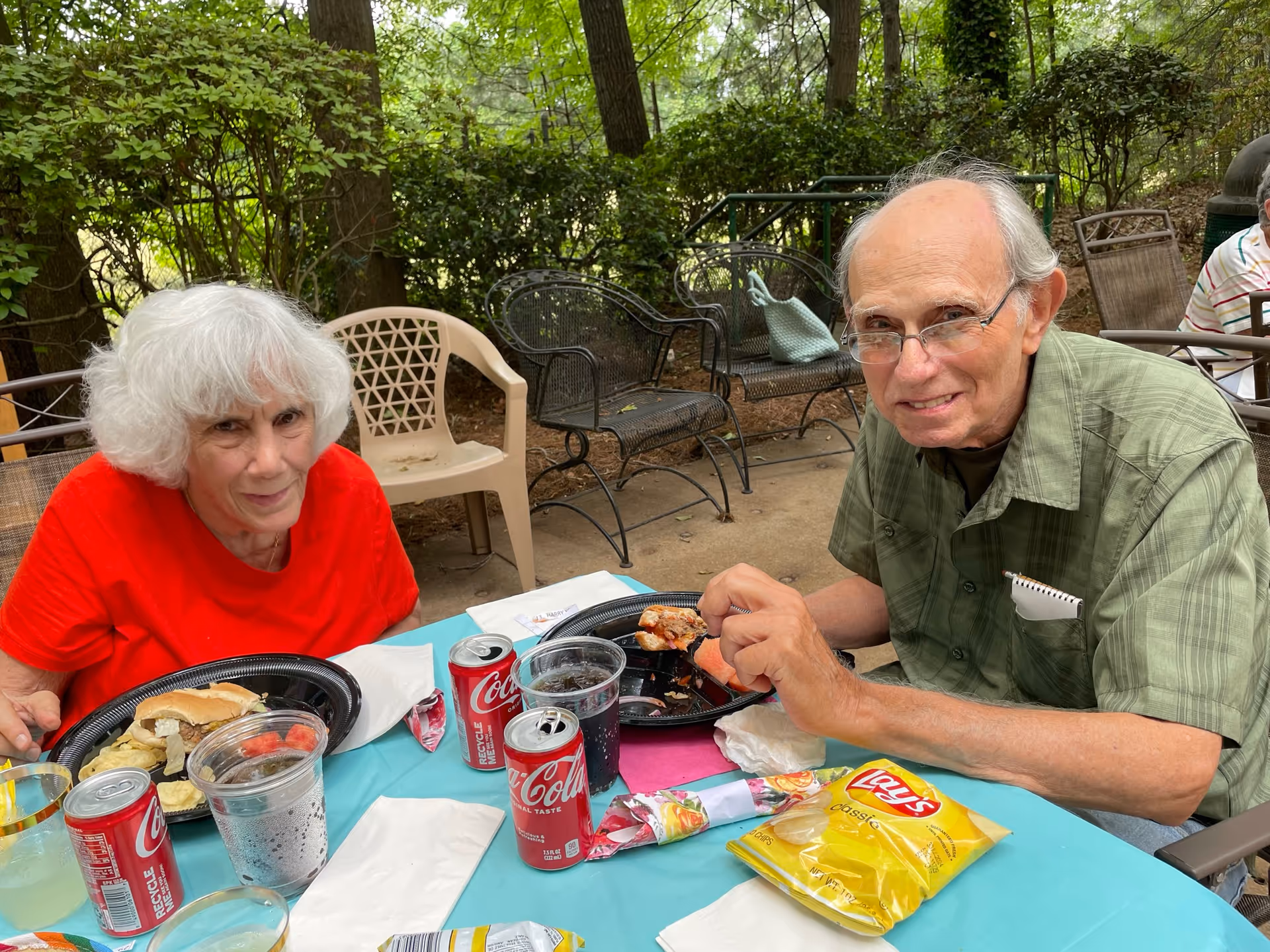 Two elderly individuals sitting at a table outdoors, enjoying a meal together. The woman on the left is wearing a red shirt and has white hair, while the man on the right is wearing glasses and a green shirt. On the table are plates with food, cans of Coca-Cola, a bag of Lay's Classic potato chips, and plastic cups. The background shows trees and outdoor chairs.