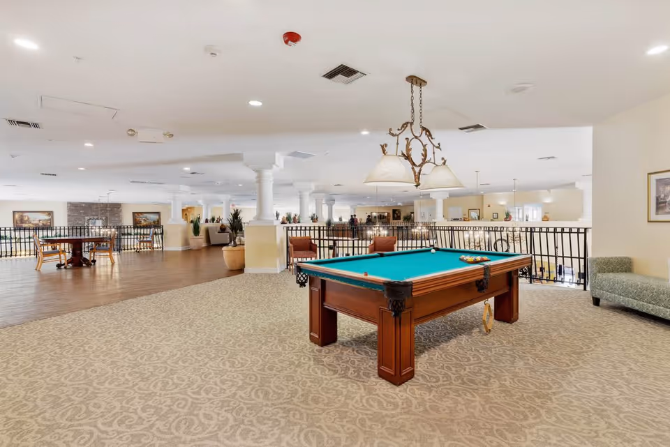 Spacious senior living common area featuring a pool table with a green felt top, a hanging light fixture above it, and a seating area with chairs and a couch. The room has carpeted and wooden flooring sections, white pillars, and framed artwork on the walls.