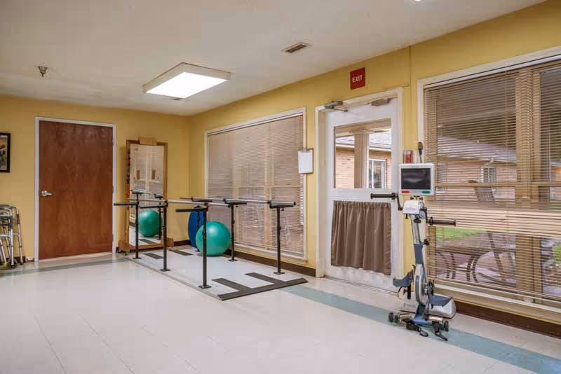 A rehabilitation room with parallel bars for walking exercises, a green exercise ball, a mirror, a walker, and a medical exercise machine. The room has large windows with blinds, a door with a small curtain, and a beige and white color scheme.