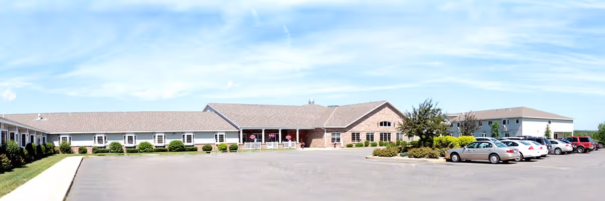 Wide exterior view of a single-story senior living facility building with a parking lot in front. The building has a combination of brick and siding with multiple windows and a covered entrance. Several cars are parked in the lot under a clear blue sky.