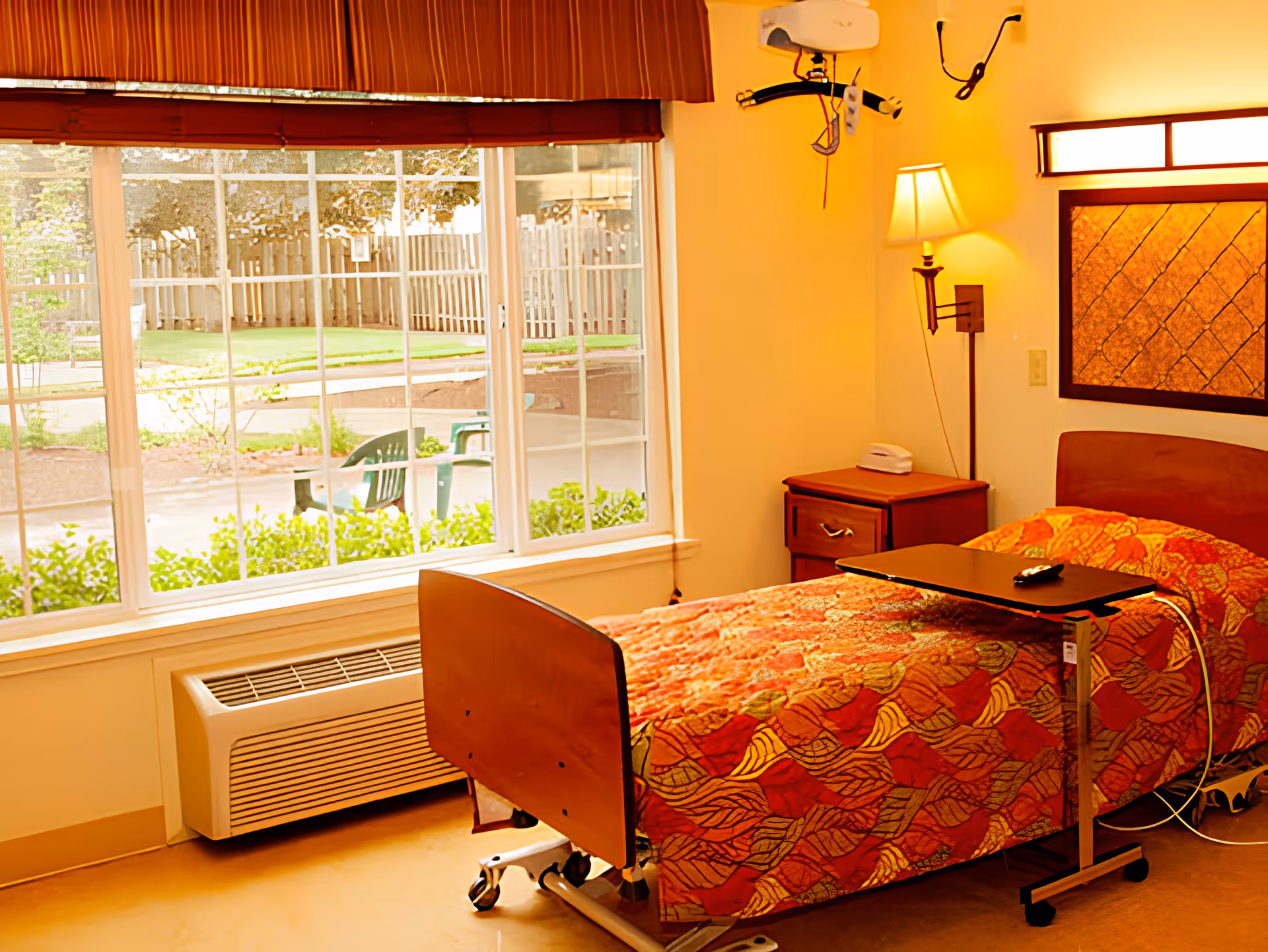 A single hospital-style bed with a colorful orange and red patterned blanket in a room with a large window showing an outdoor garden area. The room has a bedside table with a lamp and a telephone, a wall-mounted light, and a movable overbed table with a remote control on it. There is also a heating or air conditioning unit below the window.